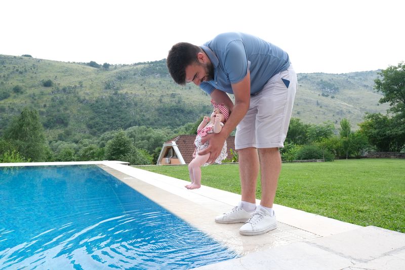 Otac drži bebu iznad bazena, planinski pejzaž u pozadini