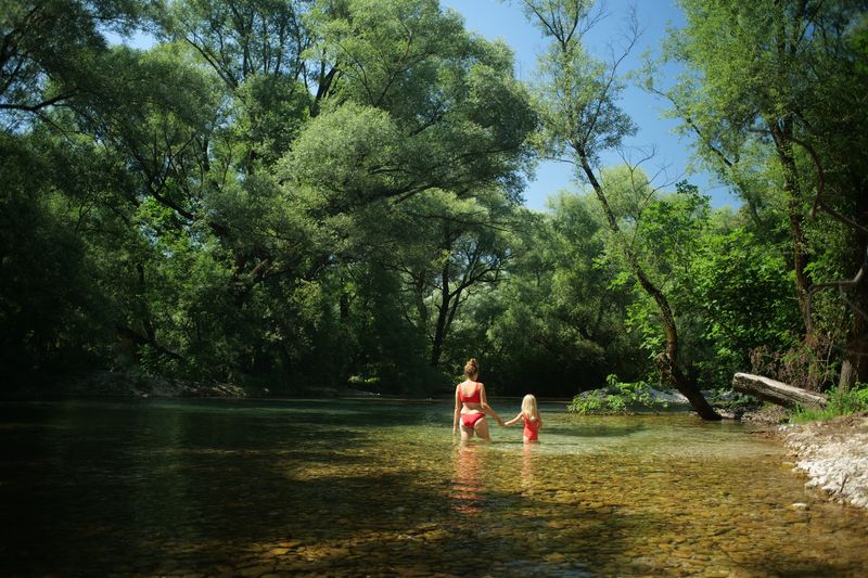 Majka i dijete gaze kroz plitku bistru rijeku okruženu drvećem